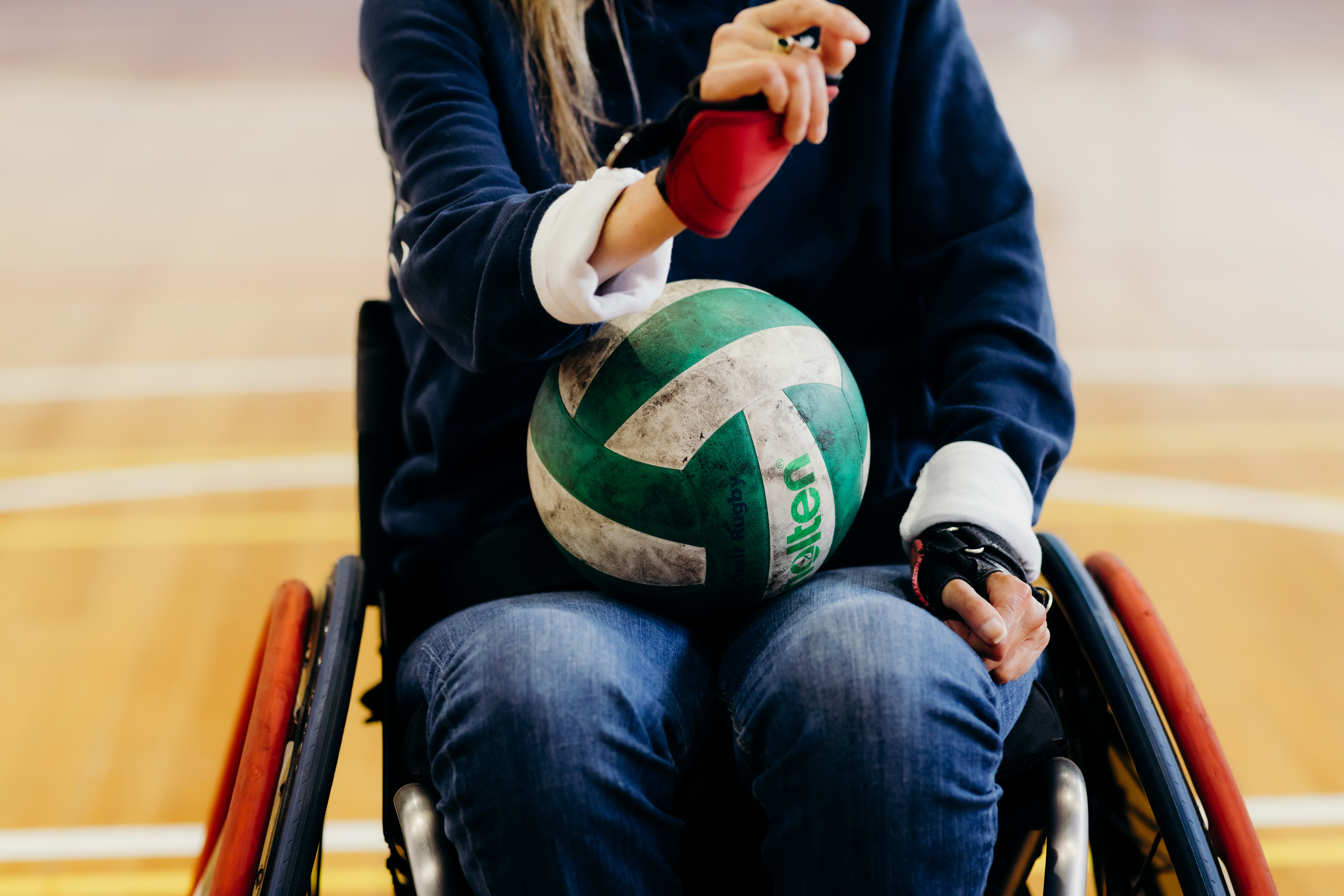 A person sitting in a wheelchair holding a green and white volleyball. The person is wearing a navy blue long-sleeve shirt, jeans, and red wrist guards. The background shows an indoor sports court with wooden flooring.