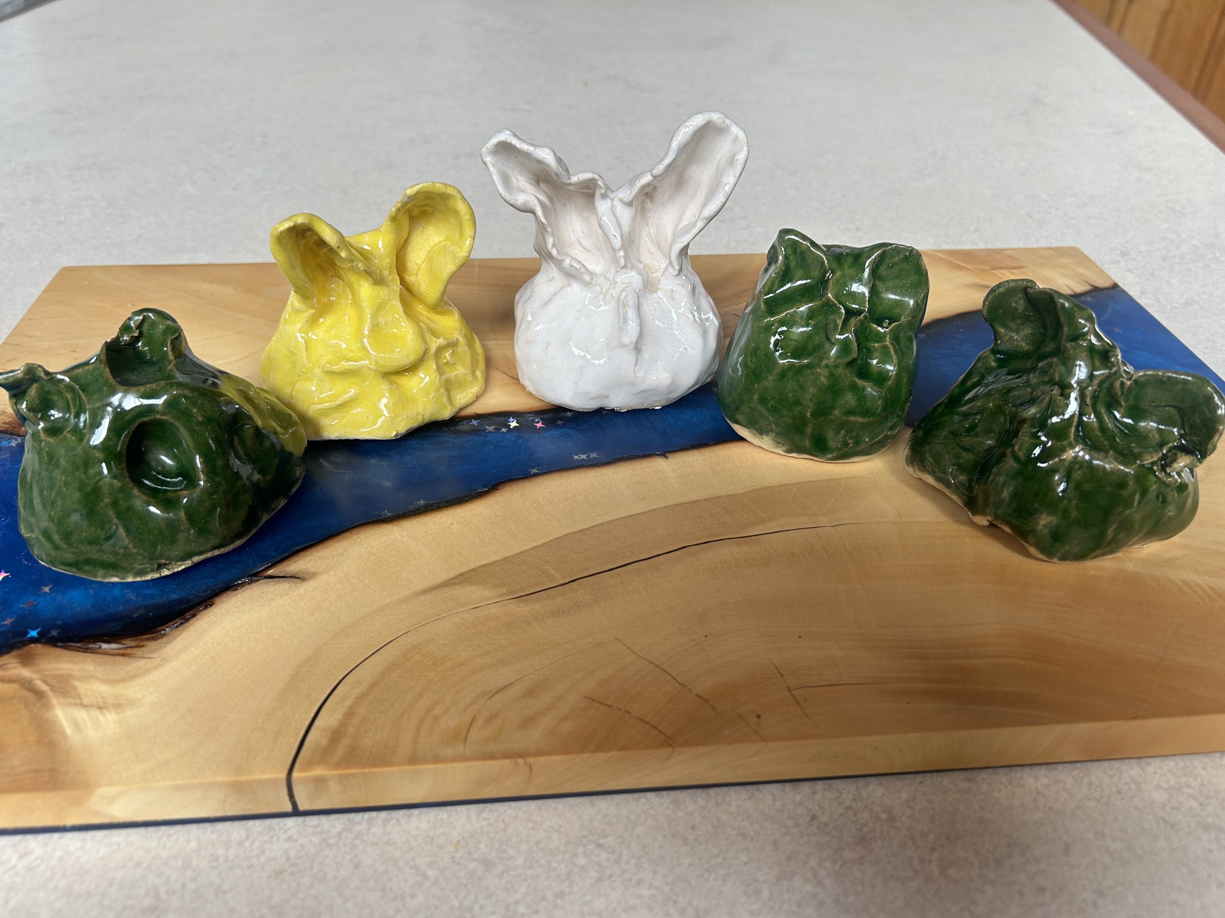 Five small clay & ceramic sculptures are positioned in a row on a wooden board. While all pieces are different they all have ears pricked up like Hannah's favourite animals - bunnies, dogs, and cats. Three of the pieces are a deep green colour, one is yellow, and the other is white.